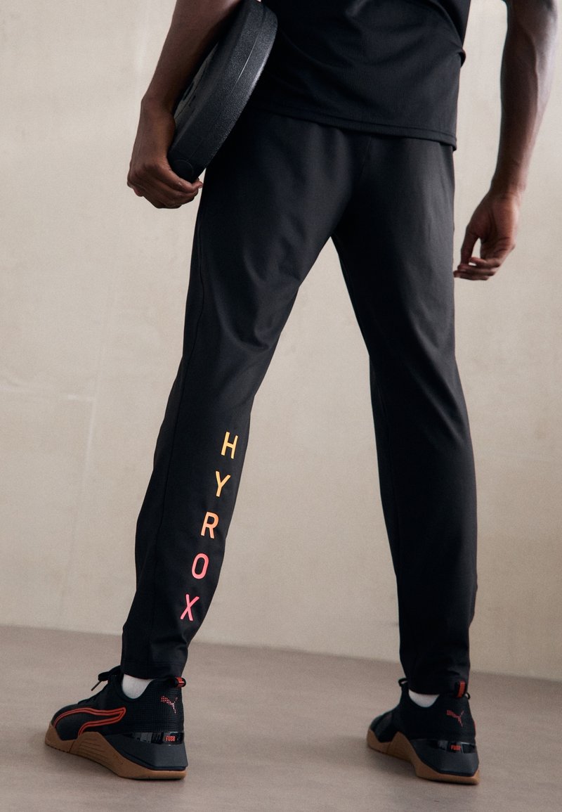 Pantalones deportivos negros con letras coloridas "HYROX" en la pierna izquierda. El material parece suave, con un diseño ajustado. Zapatillas deportivas negras con acentos rojos.