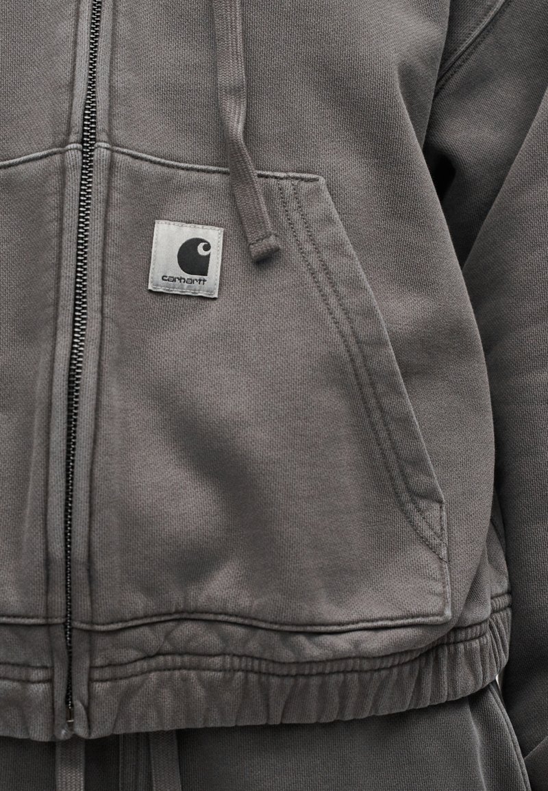 Felpa grigia con cappuccio con zip, tasca frontale, coulisse e una piccola toppa con il logo Carhartt sull'area superiore della tasca.