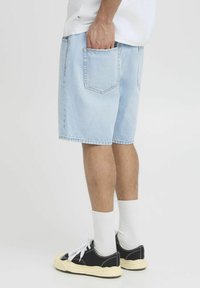 Pantalones cortos de mezclilla azul claro con corte recto y bolsillo trasero, combinados con zapatillas negras y calcetas blancas. Diseño sencillo y ajuste casual.