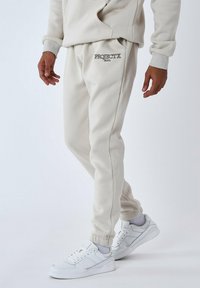 Pantalones de chándal beige en tejido suave con corte ajustado, con bolsillos laterales y el logo "PROJECT X PARIS" en gris. Combinados con zapatillas blancas.