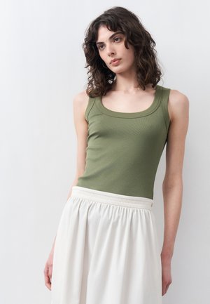 Νεαρή γυναίκα με σγουρά σκούρα μαλλιά, φορώντας ένα γιλέκο ελιάς και μια λευκή φούστα με πιέτες, στέκεται μπροστά σε απλό φόντο.