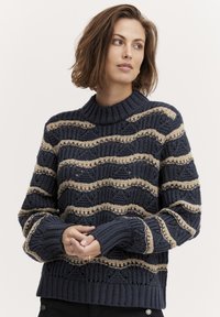 Gebreide marineblauwe trui met een textuurpatroon van beige golvende strepen, voorzien van een geribde kraag en manchetten. Grof garen met open steken.
