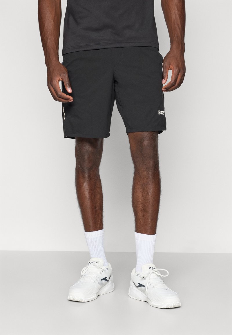 Svarta sportshorts med sidostrecker och logo på fållen. Lättviktsmaterial, elastisk midjeband, längd ovanför knäna. Bärs med vita sneakers.