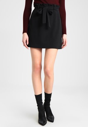 Femme portant une mini-jupe noire taille haute avec une ceinture nouée, un haut bordeaux à manches longues et des bottines noires à bout pointu.