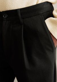 Close-up of a person wearing black pants with a buttoned waistband and a beige long-sleeve top, hand in the pants pocket.