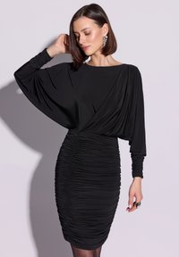 Czarna sukienka z luźnym, drapowanym topem i dopasowanym, marszczonym dołem. Długie rękawy z marszczonymi detalami. Gładka tkanina tworzy elegancką sylwetkę.