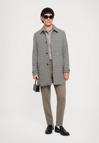 Manteau à carreaux gris et noir avec une coupe droite, porté sur une chemise claire et un pantalon marron, accessoirisé avec des lunettes de soleil noires et un petit sac.