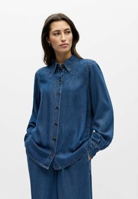 Femme portant une chemise en denim bleu avec broderies florales sur les épaules et un pantalon large en denim assorti, mains dans les poches, regardant vers la droite.