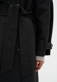 Manteau noir en trench avec une ceinture nouée, présentant un détail de bouton et un large col. La manche intérieure montre un poignet en tricot beige.
