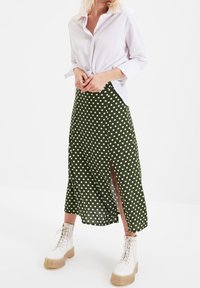 Jupe verte à pois avec une fente latérale, en tissu léger ; associée à une chemise blanche nouée et des bottes blanches robustes.