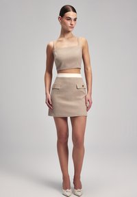 Beige geweven crop top met dunne bandjes, gecombineerd met een high-waisted mini rok met voorzakken en gouden knopen. Hakken maken de look compleet.
