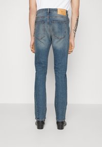 Jeans de mezclilla azul con un diseño de pierna recta, que cuentan con un parche de cuero en la parte trasera, cinco bolsillos y un ligero desgaste. Zapatos negros visibles.