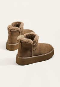 Hnedé semišové členkové čižmy s umelou kožušinou vnútri, zaoblenou špičkou a textúrovanou platformou. Obsahujú mäkké, plyšové lemy a prešitie.