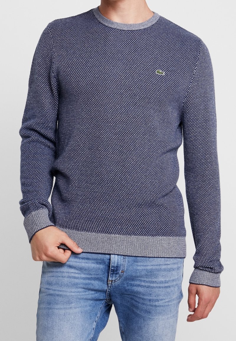 Homme portant un pull texturé bleu marine avec poignets et ourlet gris, associé à un jean bleu clair sur un fond uni.