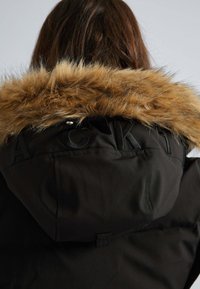 Sort parka-jakke med en hætte foret med pels. Teksturen er glat, med broderede bogstaver på ryggen og lynlåsdetaljer.