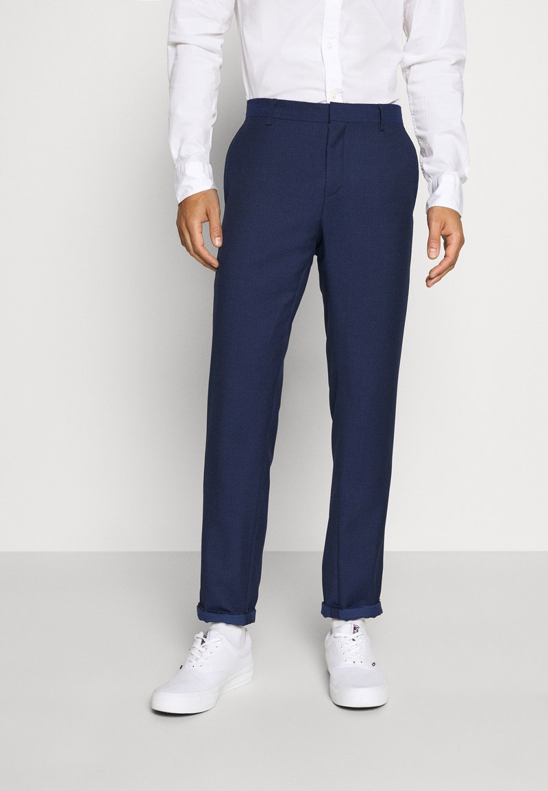 Homme portant un pantalon bleu marine sur mesure avec revers roulés, des baskets blanches et une chemise blanche boutonnée, debout sur un fond uni.