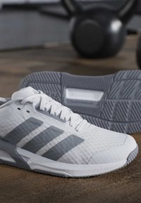 Chaussures de course Adidas blanches et grises sur un sol en bois avec un kettlebell noir flou en arrière-plan.