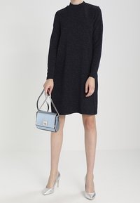 Schwarzes Kleid mit langen Ärmeln und hohem Kragen, aus strukturiertem Stoff. Kombiniert mit einer hellblauen Handtasche und silbernen High Heels.