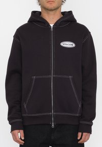 Haut à capuche zippé noir avec des accents de couture blanche et des poches avant ; comporte un logo "Volcom" brodé sur la poitrine ; tissu doux.