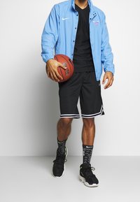 Blå lättviktig jacka med elastiska ärmar, svart t-shirt, svarta shorts med vita ränder, mönstrade svarta strumpor och svarta sneakers. Håller i en basketboll.