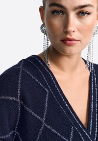 Tumesinine kootud sviiter V-kaelusega ja geomeetriliste hõbedaste niitmustritega. Mudel kannab pikki, sädelevaid kõrvarukke, millel on kaskaadsed detailid.