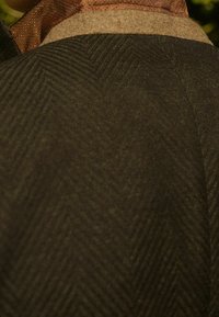 Tkanina o ciemnozielonym wzorze jodełki, z charakterystycznym teksturowanym splotem i kontrastującym brązowym kołnierzem, tworzącym warstwowy wygląd.