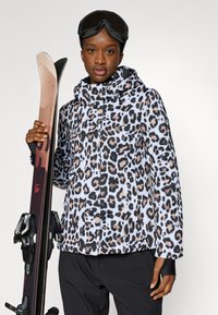 Giacca da sci con stampa leopardata in bianco, nero e beige, con cappuccio. Il modello tiene gli sci. Occhiali da sci neri completano l'outfit.