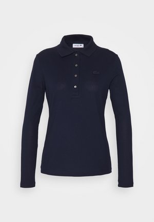 Tummansininen pitkähihainen polo paita, joka on valmistettu pehmeästä kankaasta. Paitassa on kolme nappia edessä, ribbireunainen kaulus ja pieni brodeerattu logo rinnassa.