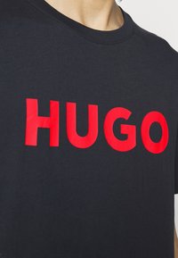 Černé bavlněné tričko s výrazným červeným textem "HUGO". Design má kulatý výstřih a krátké rukávy, s hladkou a jemnou strukturou.