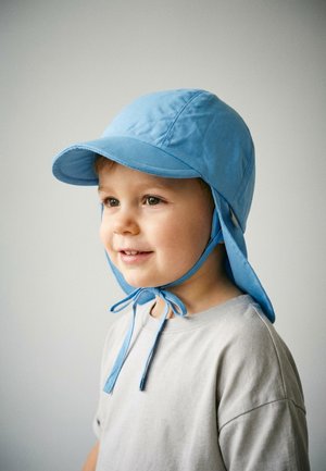 Young child smiling, wearing a light blue sun hat with chin straps tied, and a light grey shirt, facing slightly right against a pale background.