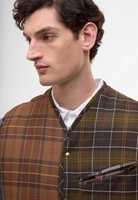 Hnědo-zelená kostkovaná vesta se zlatými knoflíky, s špičatým límcem a boční kapsou. Texturovaná vlněná látka s kontrastními vzory.