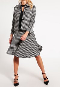 Černobílý houndstooth kabát s kulatými černými knoflíky, zkombinovaný s odpovídajícími rozšířenými sukní. Modelka nosí černé podpatky.
