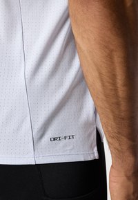 Weißes Dri-Fit Sportshirt mit perforierter Textur, seitlichem Schlitzsaum und schwarzem Logo am unteren Rand, kombiniert mit schwarzen Sportshorts.
