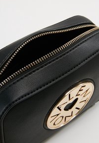 Musta nahkalaukku, jossa on kiiltävä kultainen pyöreä logo edessä ja kultaiset vetoketjut. Pinnassa on sileä pinta ja pyöristetyt muodot.