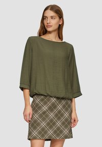 Haut texturé vert olive avec une coupe ample et des manches à poignets, associé à une jupe ajustée à carreaux dans des tons olive et blanc.