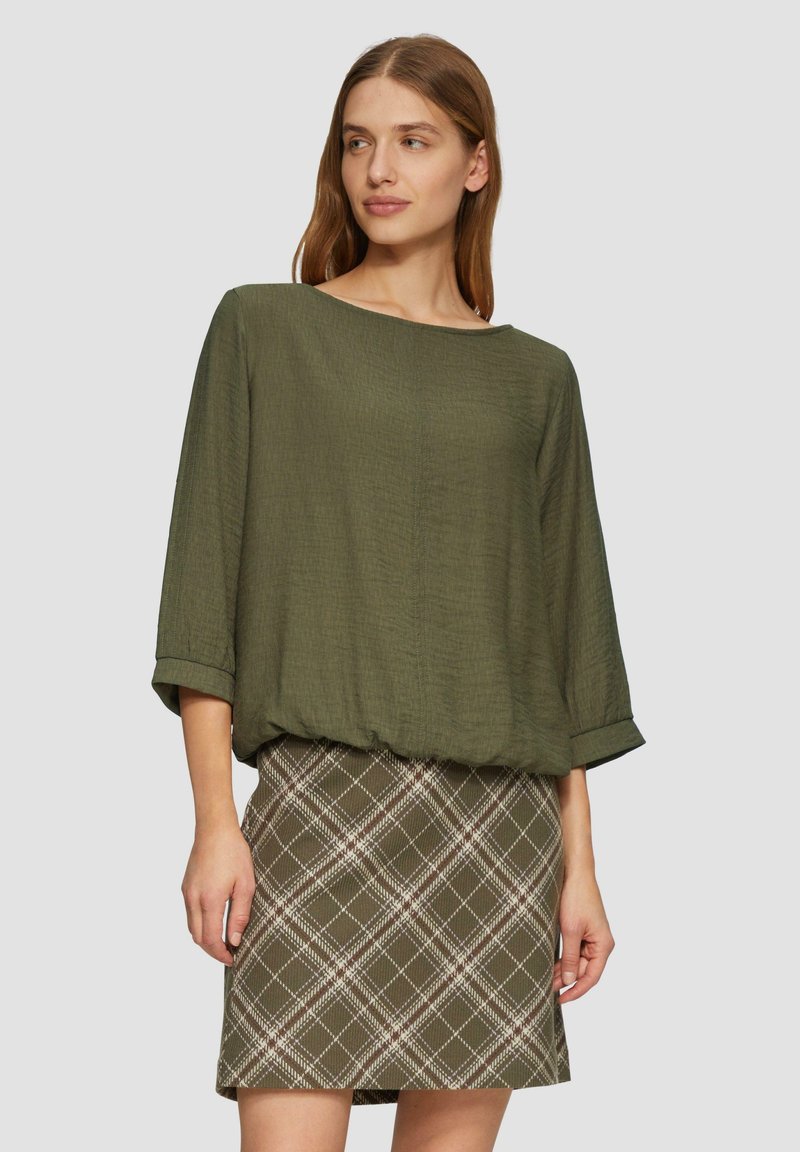 Haut texturé vert olive avec une coupe ample et des manches à poignets, associé à une jupe ajustée à carreaux dans des tons olive et blanc.