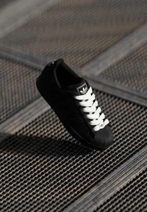 Baskets noires Adidas avec des lacets blancs flottant au-dessus d'une surface en grille métallique texturée sous un éclairage doux.