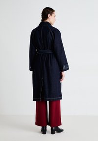 Denimjacke mit tailliertem Gürtel, dunkelblauer Farbe, Kontrastnähten, langen Ärmeln und seitlichen Schlitzen. Kombiniert mit weiten roten Hosen und Ankle Boots.