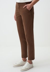 Pantaloni di cotone marroni con gamba dritta, tasche laterali e orli cuffed, abbinati a sneaker bianche. Texture liscia e design sobrio.
