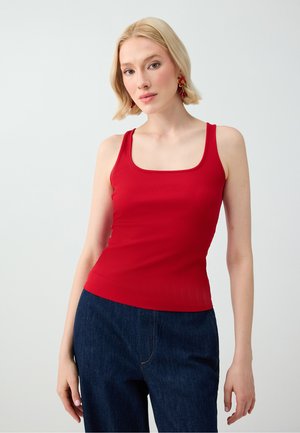 Jeune femme aux cheveux blonds courts, portant un haut rouge sans manches, un jean bleu foncé et des boucles d'oreilles rouges ornées de fleurs, debout devant un fond uni et clair.