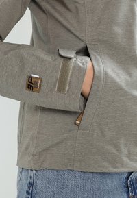 Veste à manches longues gris clair, dotée d'une fermeture velcro aux poignets, d'une poche visible et d'une étiquette de marque sur la manche. Tissu doux et texturé.