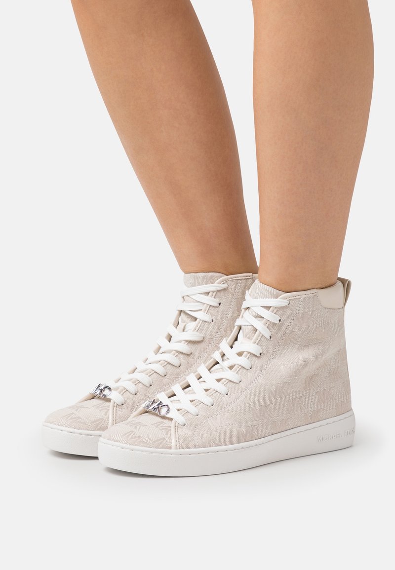 Beige-kankaiset korkeat tennarit, joissa on teksturoitu MK-logokuosi, valkoiset nauhat ja kumipohja. Varressa on hopeista metalliosaa.