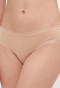 Nude-farget bikini-truse laget av glatt stoff, med en delikat bølget blondekant og minimal dekning.
