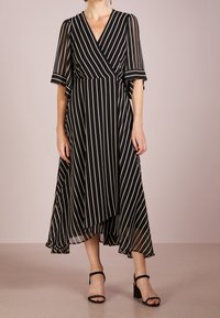 Raitapaitainen mekko mustavalkoisena, jossa on läpinäkyvät hihat, V-pääntie ja virtaava helma. Sisältää kietaisumallin sekä nilkkapituuden.