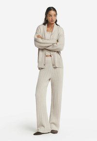 Lichtbeige gebreide cardigan met een sjaalkraag, gecombineerd met bijpassende brede kabelgebreide broek. Gladde textuur, casual pasvorm en subtiele details.