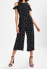Combinaison noire à pois avec design épaules dénudées, manches à volants, jambes larges et pois de différentes couleurs en rose et blanc.
