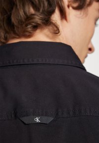 Camisa negra con cuello puntiagudo y textura suave. Presenta un pequeño parche con el logo blanco "ck" en la parte posterior del cuello. Diseño simple y moderno.