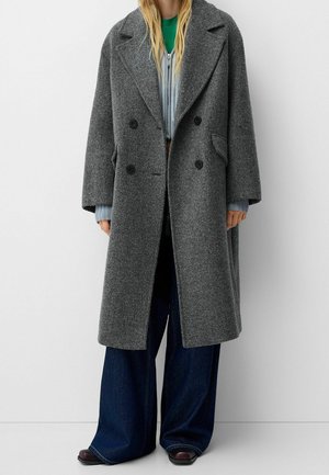 Personne portant un long manteau en laine gris à double boutonnage, un haut vert, un pull à fermeture éclair bleu clair, un jean large bleu foncé et des chaussures noires.