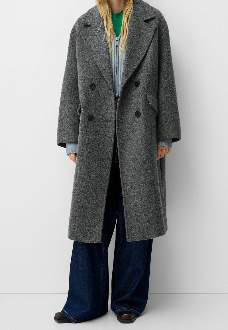 Personne portant un long manteau en laine gris à double boutonnage, un haut vert, un pull à fermeture éclair bleu clair, un jean large bleu foncé et des chaussures noires.