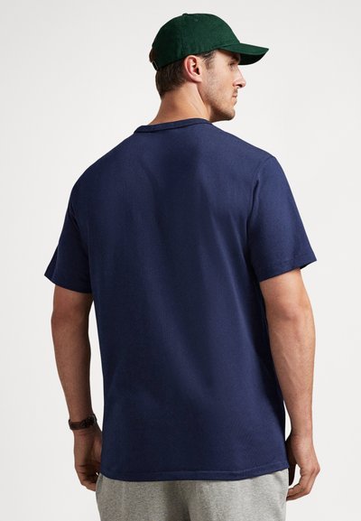 Moški, ki nosi temno zeleno kapo, mornarsko modro kratko majico, sive hlače in ročno uro, obrnjen stran na enobarvni beli podlagi.